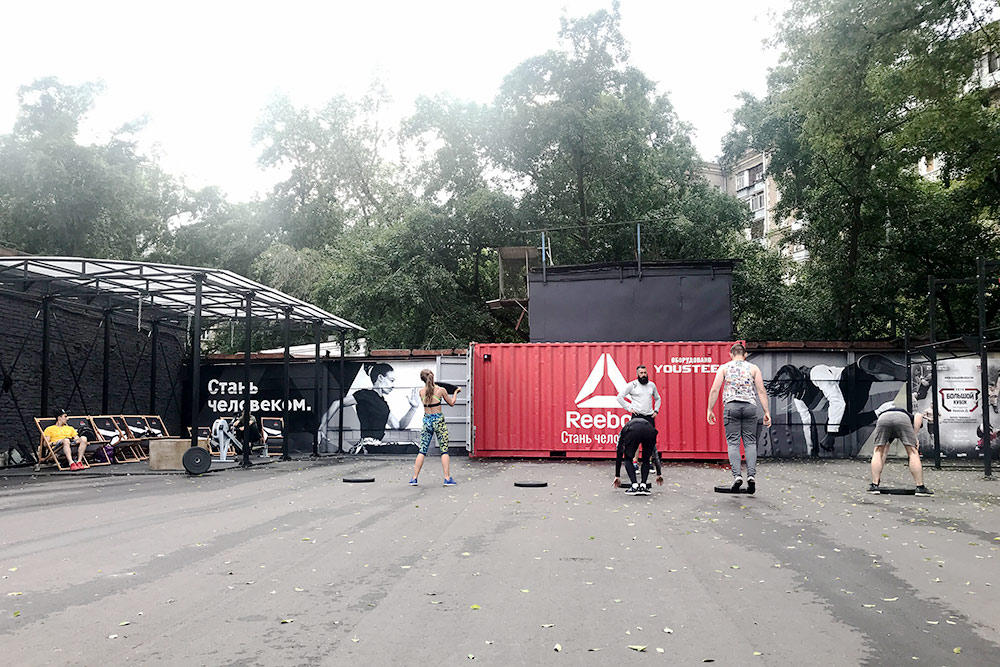По программе «Рибок в парках» мы тренировались в городском пространстве «Хлебозавод». На площадке — турники и небольшой бокс со штангами, блинами, гребным тренажером. На асфальт приходится падать и биться о него кроссовками — новую форму лучше не надевать. Во время пробежки приходится обегать людей, рестораны, парковку